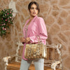 Woman in pink blazer and light blue denim jeans looking to the right and posing while holding a Nicole Lee Calista handbag in metallic gold with gold-tone hardware details. The woman is carrying it by the top handles around her arms. Set against a modern, rustic background with a bench and flowers hanging behind her. 