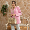 Woman in pink blazer and light blue denim jeans looking to the right and posing while holding a Nicole Lee Calista handbag in metallic gold with gold-tone hardware details. The woman is carrying it by the top handles to show the dual handles and comfortable carry. Set against a modern, rustic background with a bench and flowers hanging behind her. 