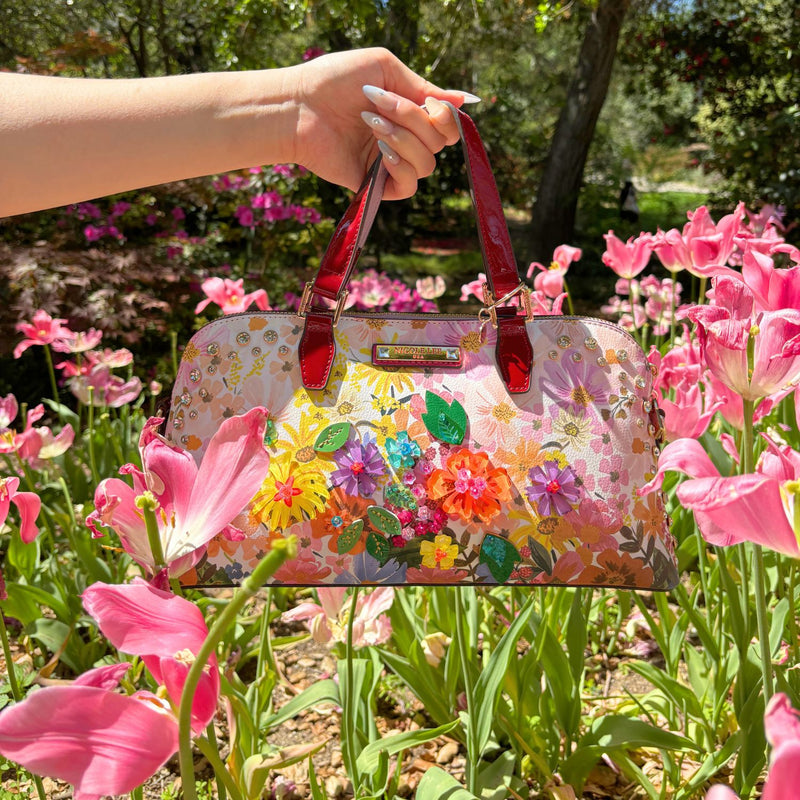 HOLDING OUR NICOLE LEE DOME HANDBAG IN DREAMS BLOSSOM DESIGN SET IN A GARDEN FULL OF PINK FLOWERS BACKGROUND.