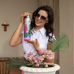 woman in white crop top and pink pants with pink ombre sunglasses smiling and holding a stainless steel bluetooth speaker tumbler with a top handle and a pink removable bluetooth speaker base in the Nicole Lee Violet Blossom exclusive fashion print in a modern background with a pot of flowers and foliage in front of her.