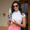woman in white crop top and pink pants with pink ombre sunglasses smiling and holding a stainless steel bluetooth speaker tumbler with a top handle and a pink removable bluetooth speaker base in the Nicole Lee Violet Blossom exclusive fashion print. 