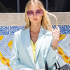 woman in sky blue long sleeve top and stylishly posing and showing off our oversized pink sunglasses set on a contemporary wall.