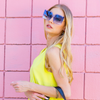 woman in yellow top and stylishly posing and showing off our oversized blue sunglasses set on a pink wall.