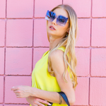 woman in yellow top and stylishly posing and showing off our oversized blue sunglasses set on a pink wall.