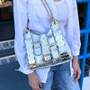 Cropped shot of a woman in white button up and blue denim jeans outside wearing the Nicole Lee Calista shoulder bag in metallic silver against a blue background.
